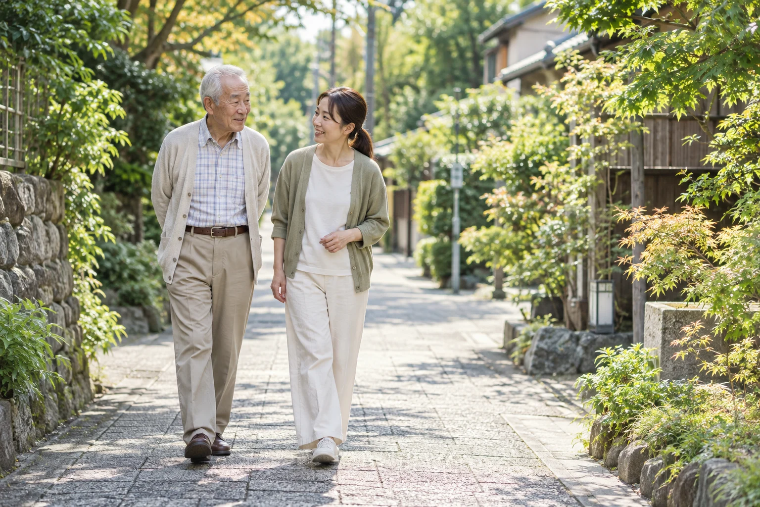 緑のある歩道を、高齢の男性と介護スタッフが一緒に歩くイメージ